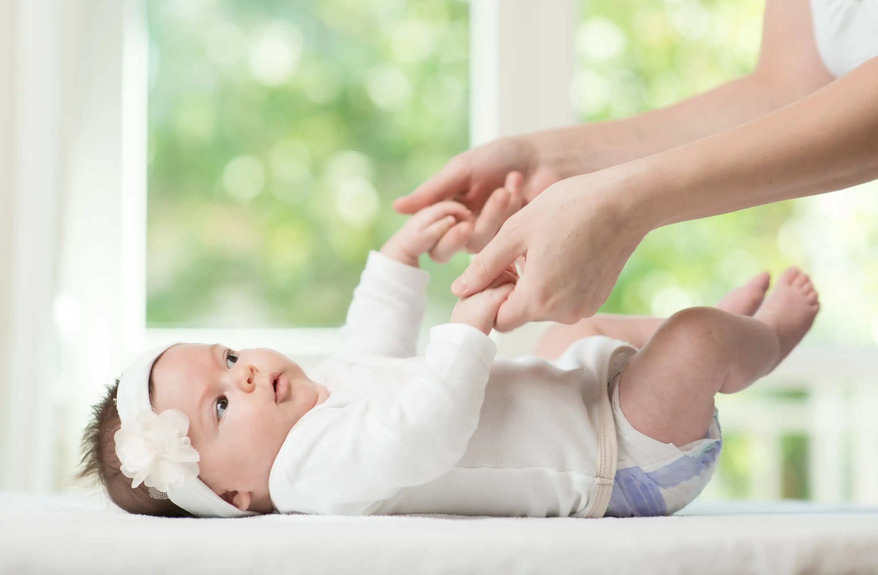 A baby wearing a white onesie and headband lies on a soft surface, looking up and holding an adult's hands.