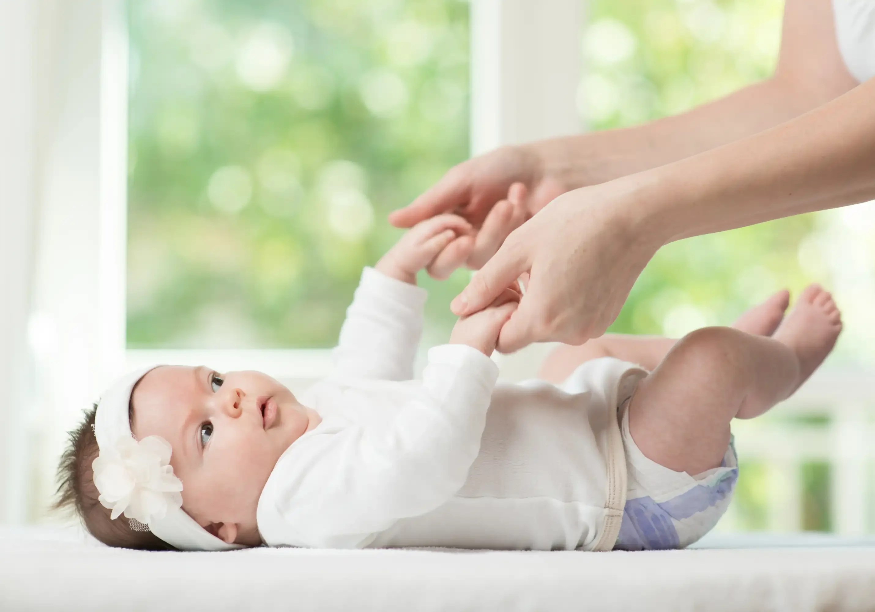 A baby wearing a white onesie and headband lies on a soft surface, looking up and holding an adult's hands.